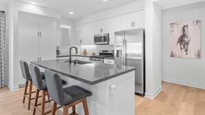 A modern kitchen with a central island, bar stools, stainless steel appliances, and white cabinetry. - Desert Hideaway: 2BR/2BA, BBQ, FirePit, PGA West! (La Quinta)
