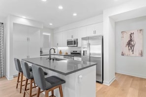 A modern kitchen with a central island, bar stools, stainless steel appliances, and white cabinetry. - Desert Hideaway: 2BR/2BA, BBQ, FirePit, PGA West! (La Quinta)