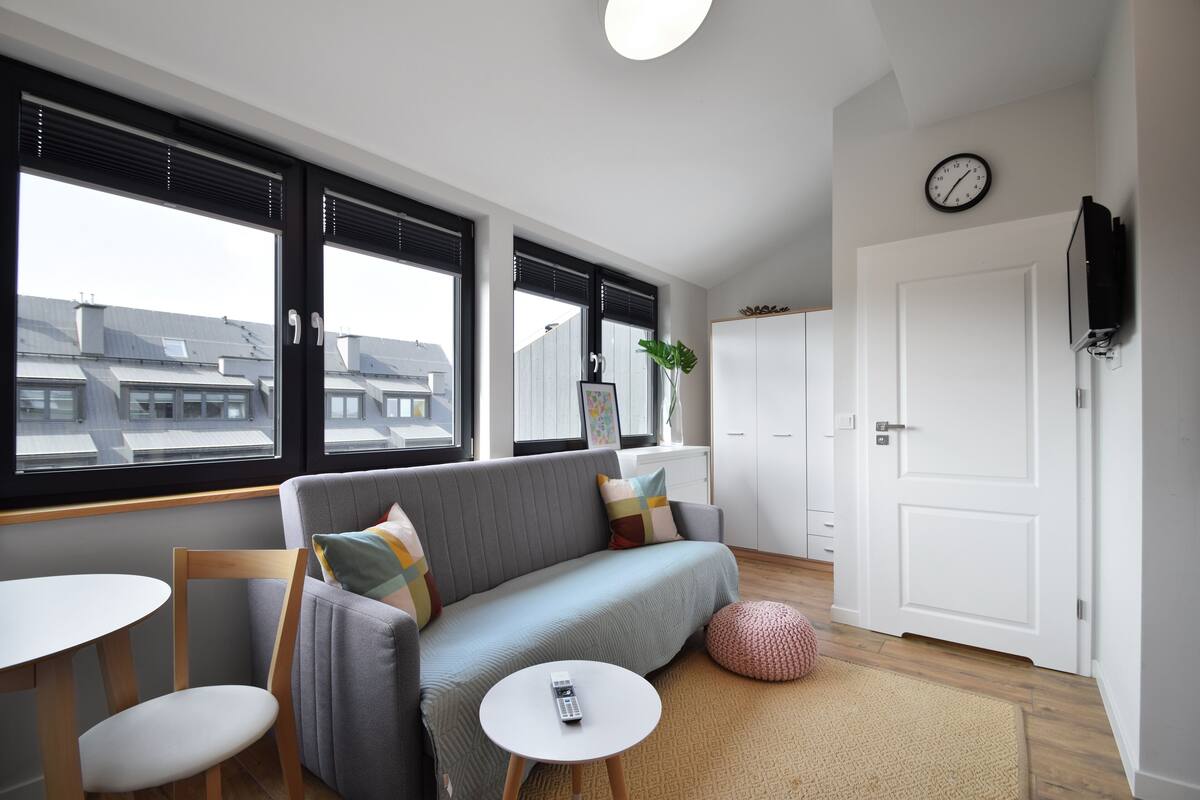 A modern living room with a grey sofa, a round white table, and a small round ottoman. There is a large window with blinds, a white door, and a wall-mounted TV.