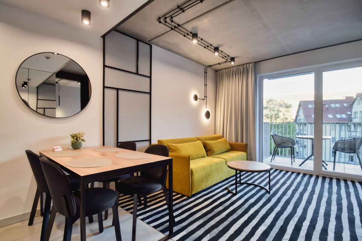 A modern living room with a dining table, sofa, and chairs, a large mirror, and a view through a sliding glass door.