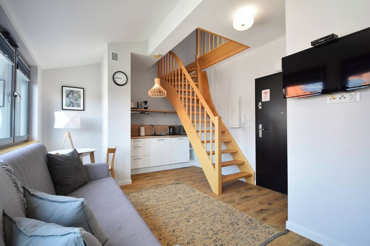 A modern living space with a wooden staircase, a grey sofa, a kitchen area, and a black refrigerator.
