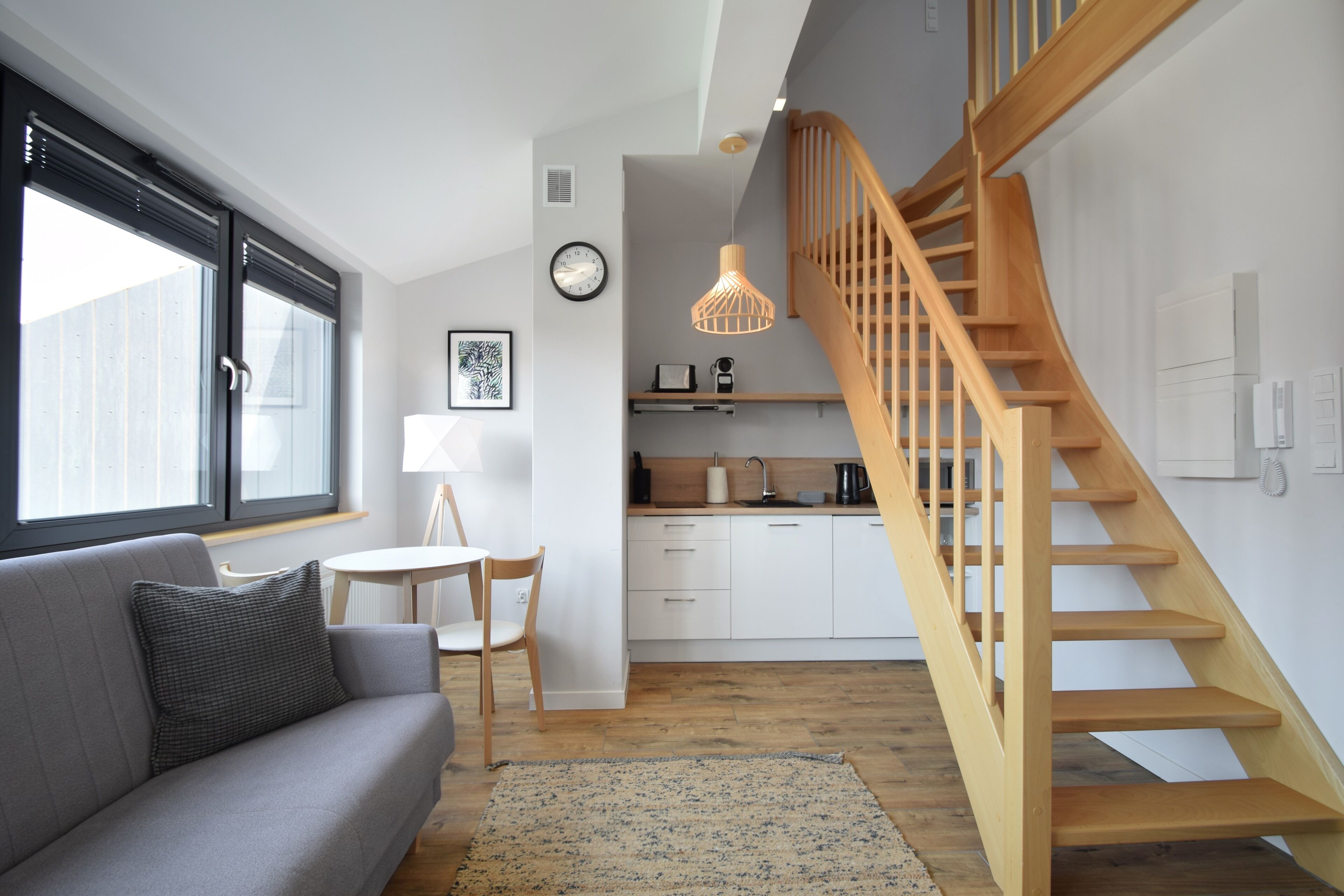 Una sala de estar moderna con un sofá gris, una escalera de madera y una zona de cocina al fondo.
