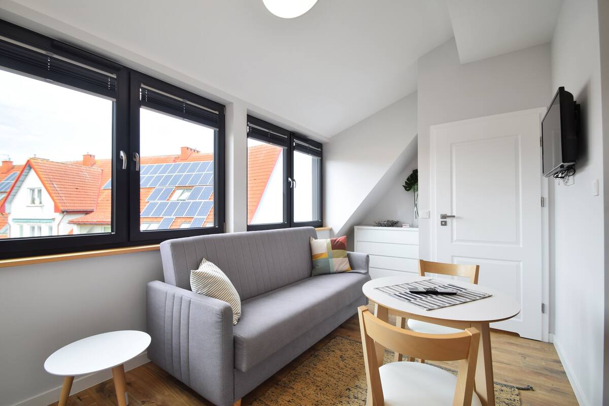 A modern living room with a grey sofa, a small white table, and a dining area with a white table and chairs. Large windows provide views of solar panels on neighboring rooftops.