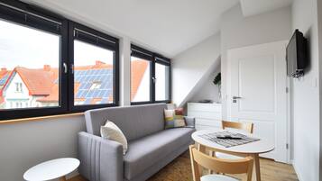 Un moderno salón con un sofá gris, una mesita blanca y un comedor con una mesa y sillas blancas. Amplias ventanas ofrecen vistas de paneles solares en los tejados de los edificios vecinos.