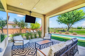 A covered patio with outdoor furniture, a flat-screen TV, and a view of a swimming pool and greenery. - Desert Chill: Pool & Spa at PGA West Stadium (La Quinta)