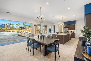 Une cuisine moderne dotée d’un îlot central, d’un coin repas avec une grande table et des chaises, et offrant une vue sur une piscine et les montagnes.