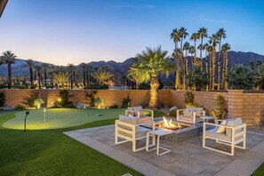 An outdoor patio with a fire pit and seating, a putting green, and palm trees.