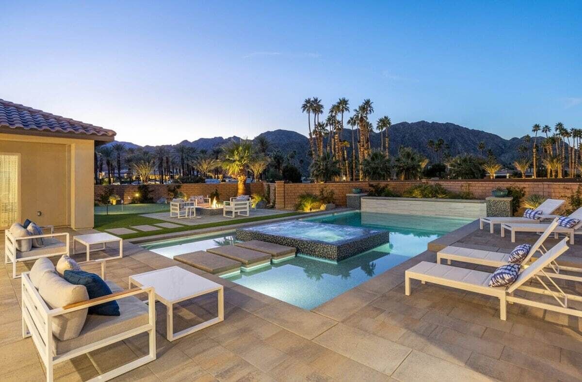 A pool area with lounge chairs and a hot tub, surrounded by palm trees and mountains.
