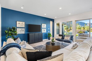 A modern living room with a blue accent wall, a flat-screen TV, and a large window offering a view of palm trees and mountains. - Poolside Paradise | Fire Pit & Golf Next Door (La Quinta)