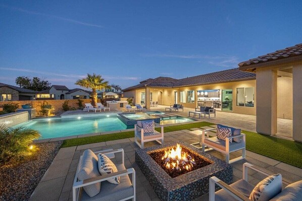 A residential outdoor area with a swimming pool, lounge chairs, a fire pit, and a patio with a view of the house.