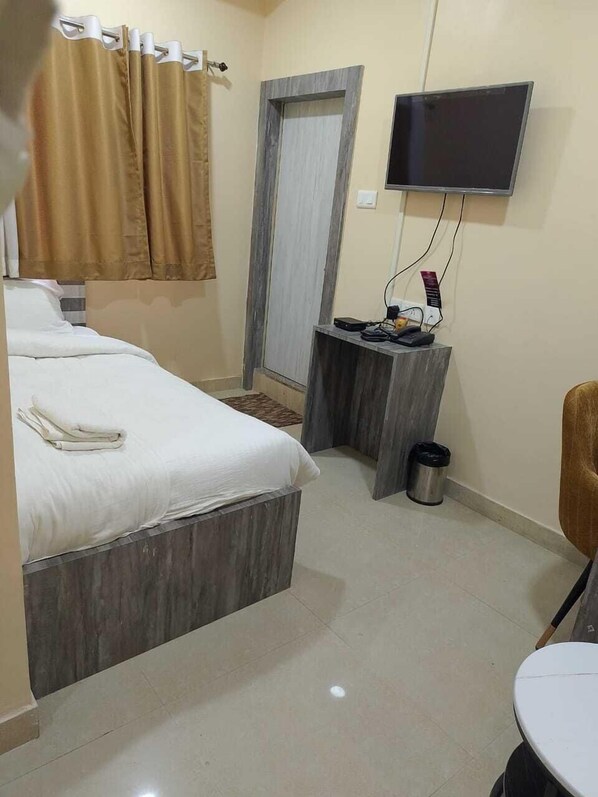 A hotel room with a bed, a small desk with a TV, a trash bin, and a chair.
