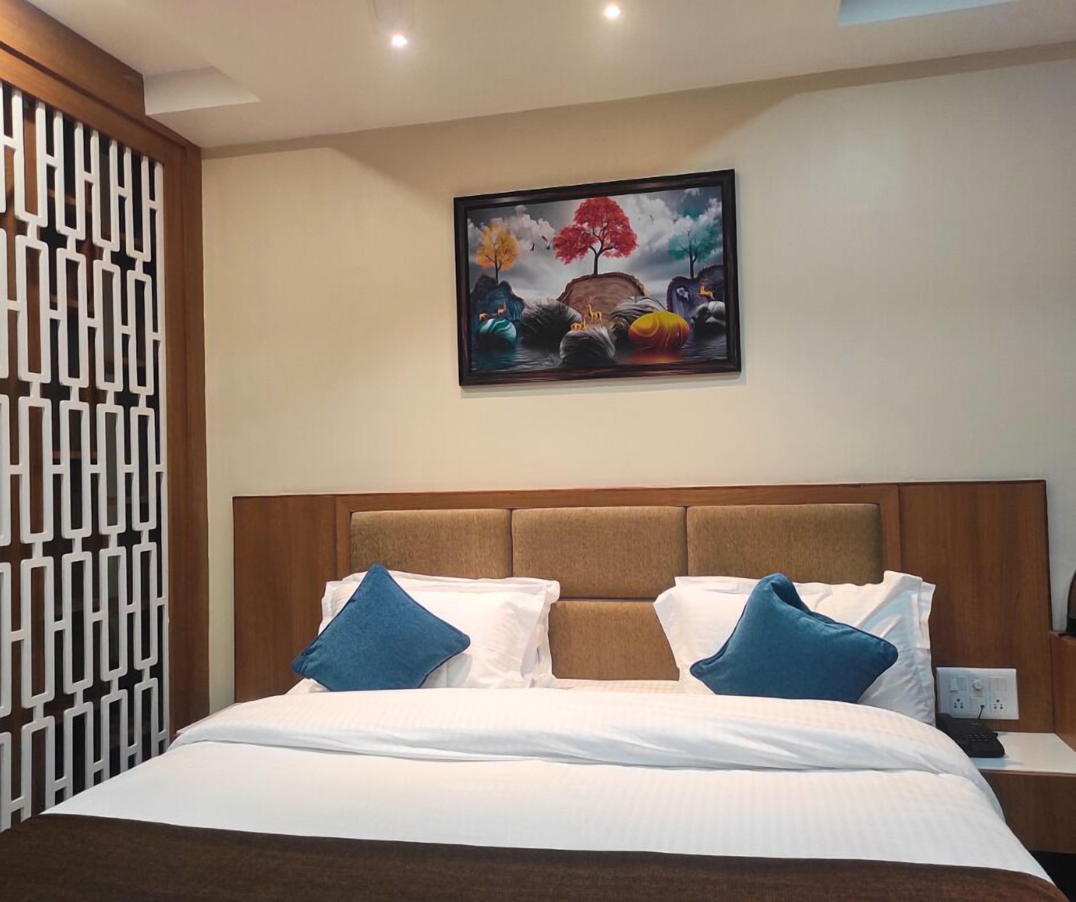 A hotel room with a bed, a headboard, a nightstand, a power outlet, and a painting on the wall.