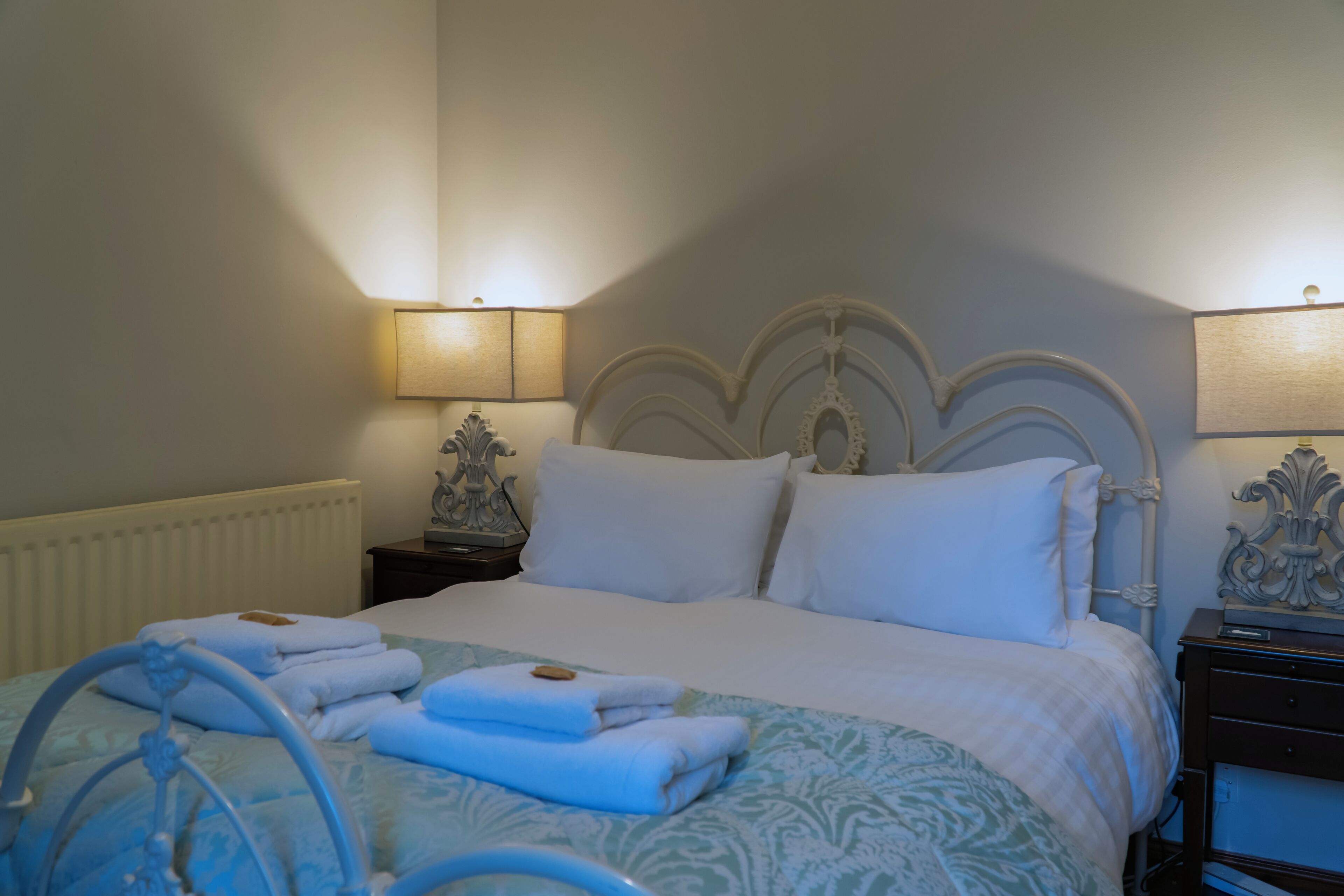A neatly made bed with white linens, a blue patterned bedspread, and folded towels on the side. There are two bedside tables with lamps, and a radiator under the bed.