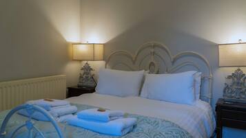 A neatly made bed with white linens, a blue patterned bedspread, and folded towels on the side. There are two bedside tables with lamps, and a radiator under the bed.