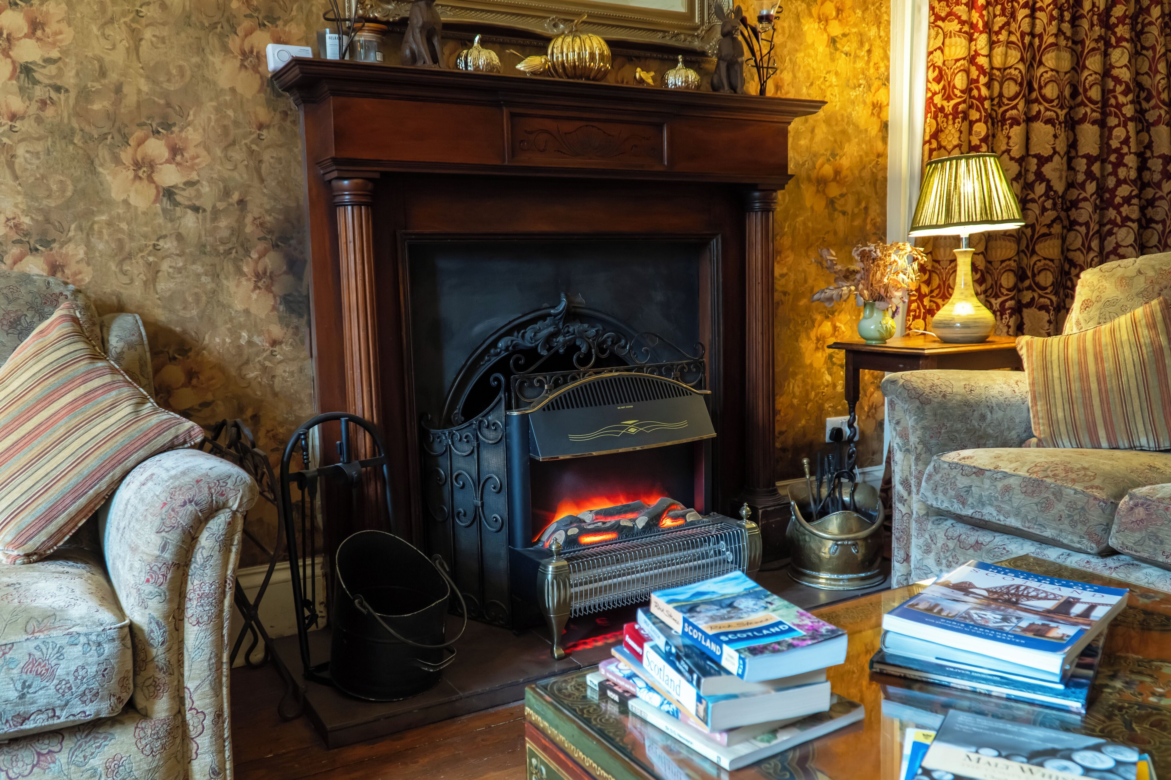 Een knusse woonkamer met een open haard, een bank, een bijzettafeltje met boeken en een lamp.