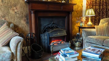 Een knusse woonkamer met een open haard, een bank, een bijzettafeltje met boeken en een lamp.