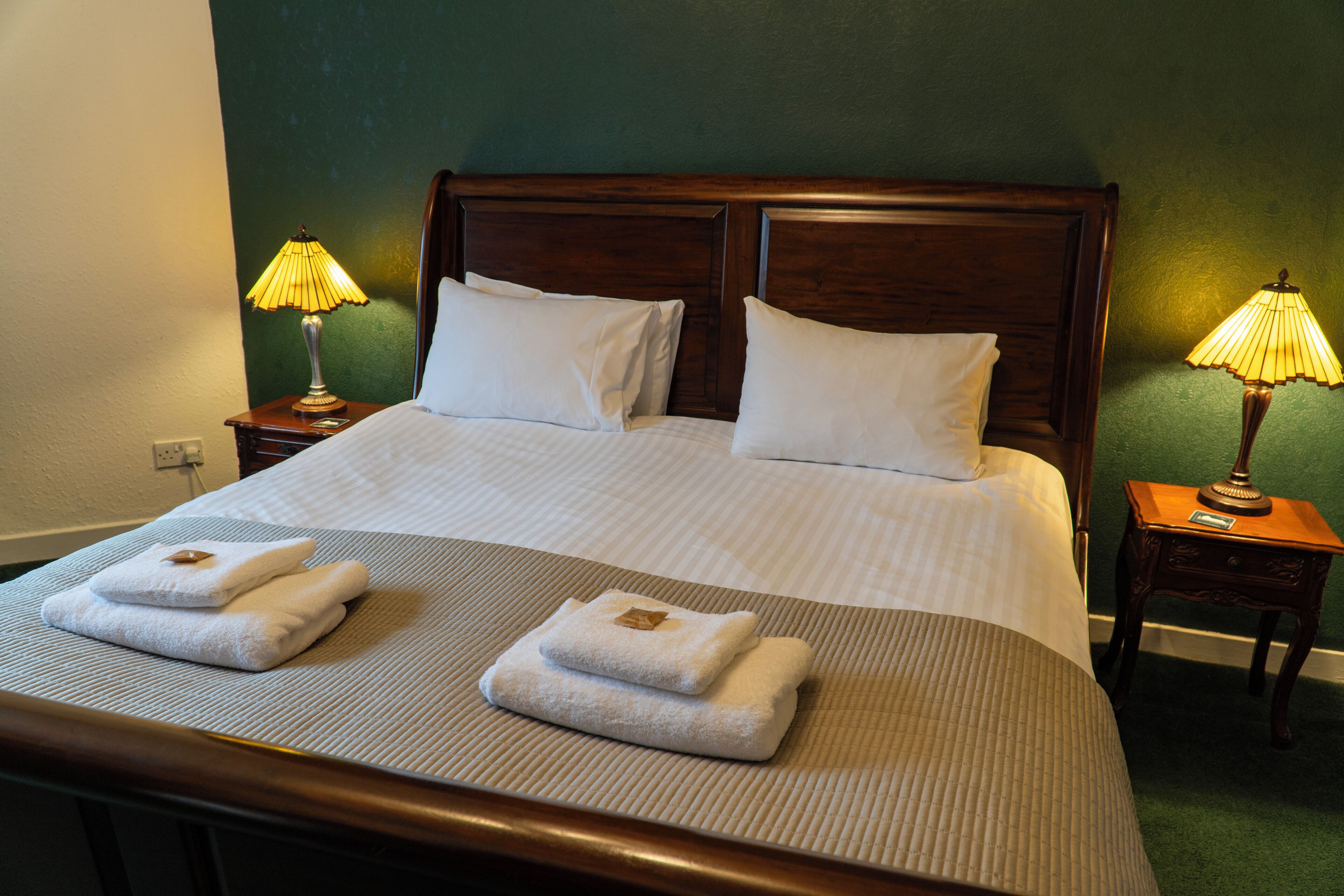 Un lit bien fait, avec du linge de lit blanc et deux oreillers, deux tables de chevet avec des lampes, et deux serviettes pliées posées sur le lit.