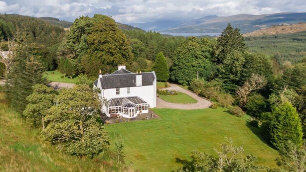 A white house with a glass conservatory, surrounded by a well-maintained lawn and dense trees. - Killean Farmhouse (Inveraray)