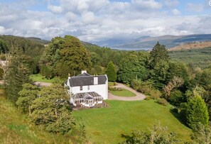 A white house with a glass conservatory, surrounded by a well-maintained lawn and dense trees. - Killean Farmhouse (Inveraray)