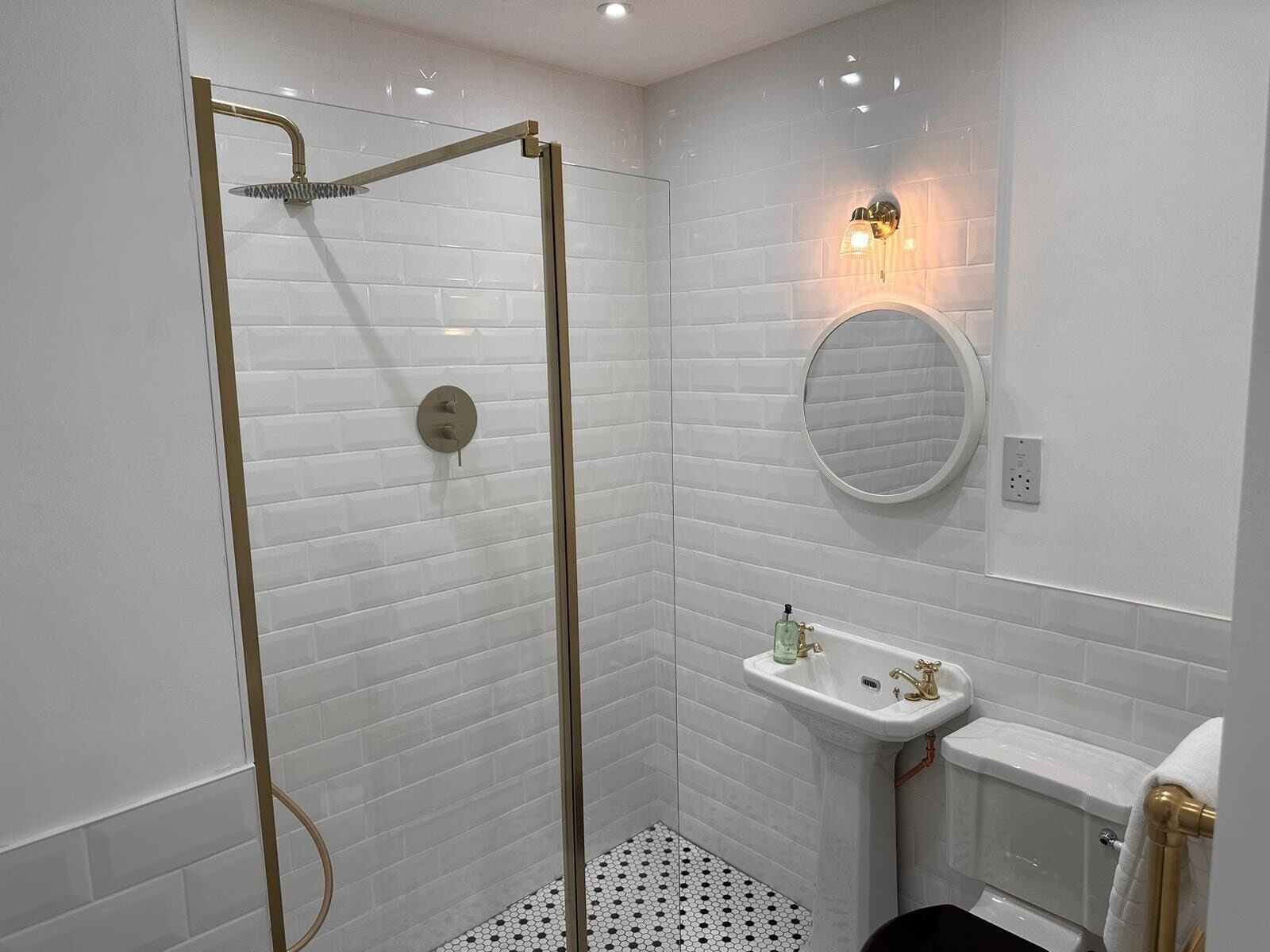 Un baño moderno con azulejos blancos tipo metro, ducha con cerramiento de vidrio, lavamanos pedestal y espejo redondo.
