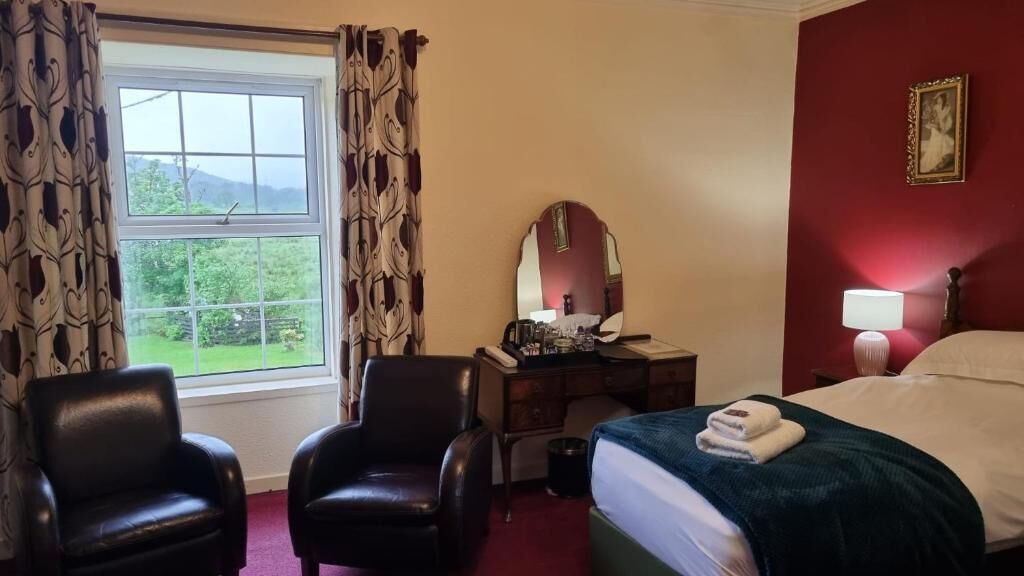 A hotel room with a bed, two chairs, a desk with a mirror, a window with curtains, and a red wall with a framed picture.