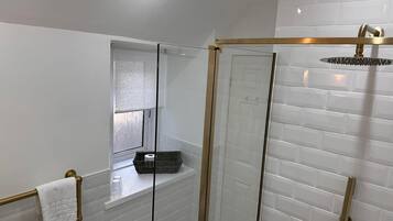 Un baño moderno con azulejos blancos tipo metro, una ducha con cerramiento de vidrio, un cabezal de ducha dorado y una ventana con persiana de vidrio esmerilado.