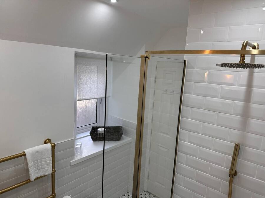 Un baño moderno con azulejos blancos tipo metro, una ducha con cerramiento de vidrio, un cabezal de ducha dorado y una ventana con persiana de vidrio esmerilado.