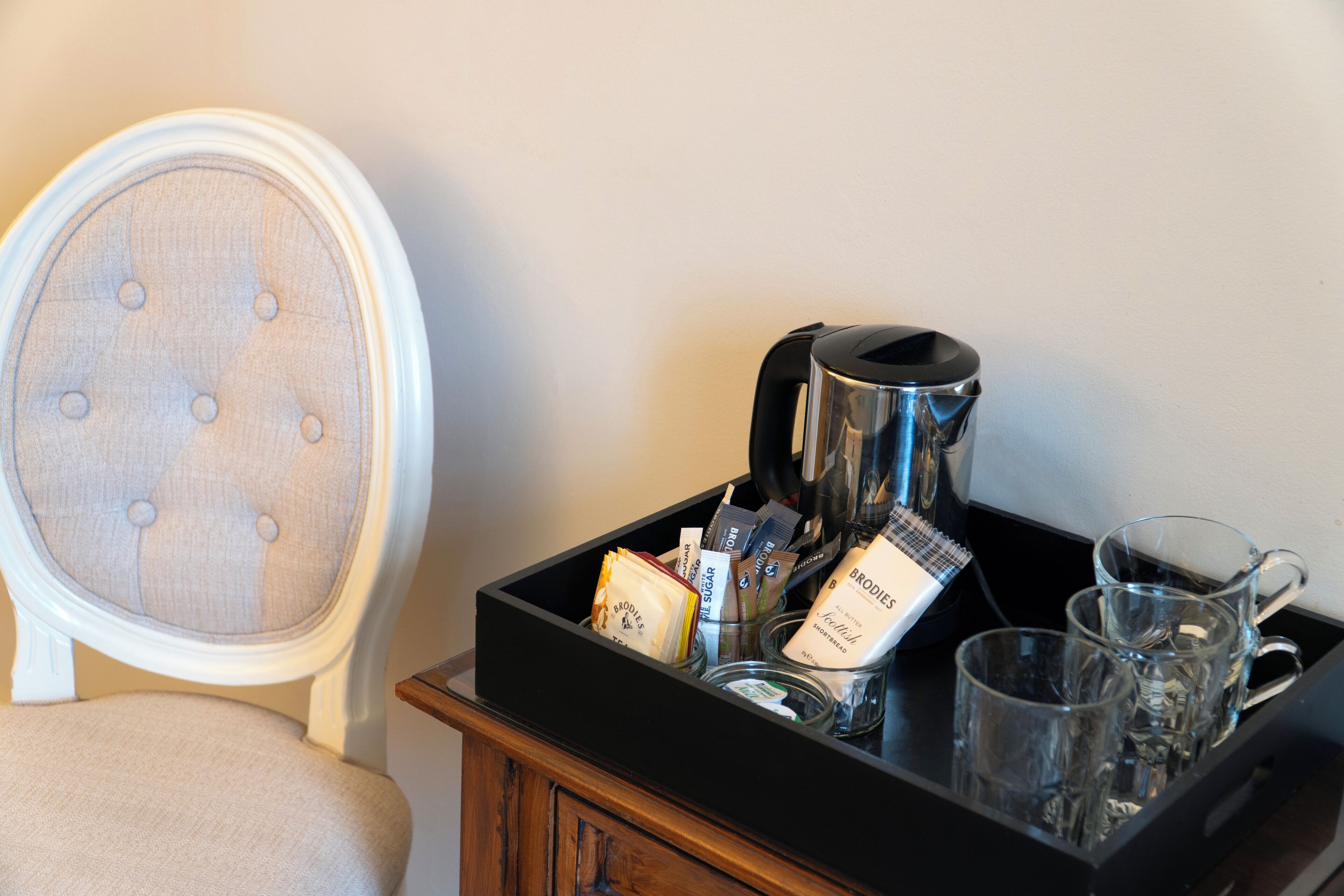 A tray with a coffee maker, packets, and glasses on a wooden table next to a chair.