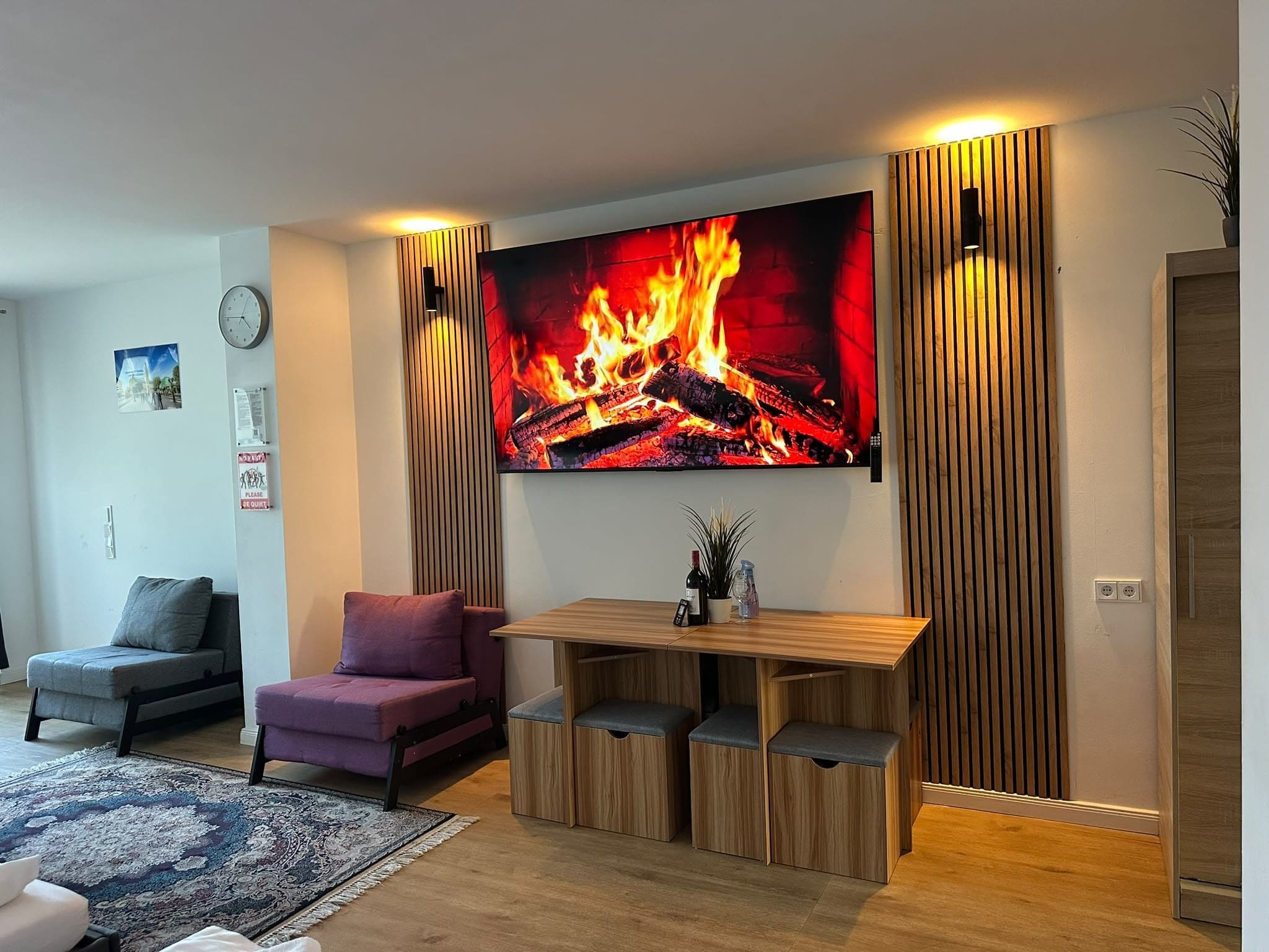 Ein modernes Wohnzimmer mit einem großen Flachbildfernseher, der ein Bild eines Kaminfeuers zeigt, einer Couch, einem kleinen Tisch mit einer Weinflasche und einer Zierpflanze.