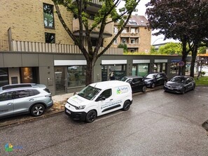 A white van parked in front of a building with a glass facade, a tree in the foreground, and a parked car to the left.
