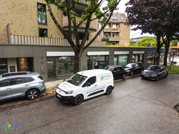 A white van parked in front of a building with a glass facade, a tree in the foreground, and a parked car to the left.