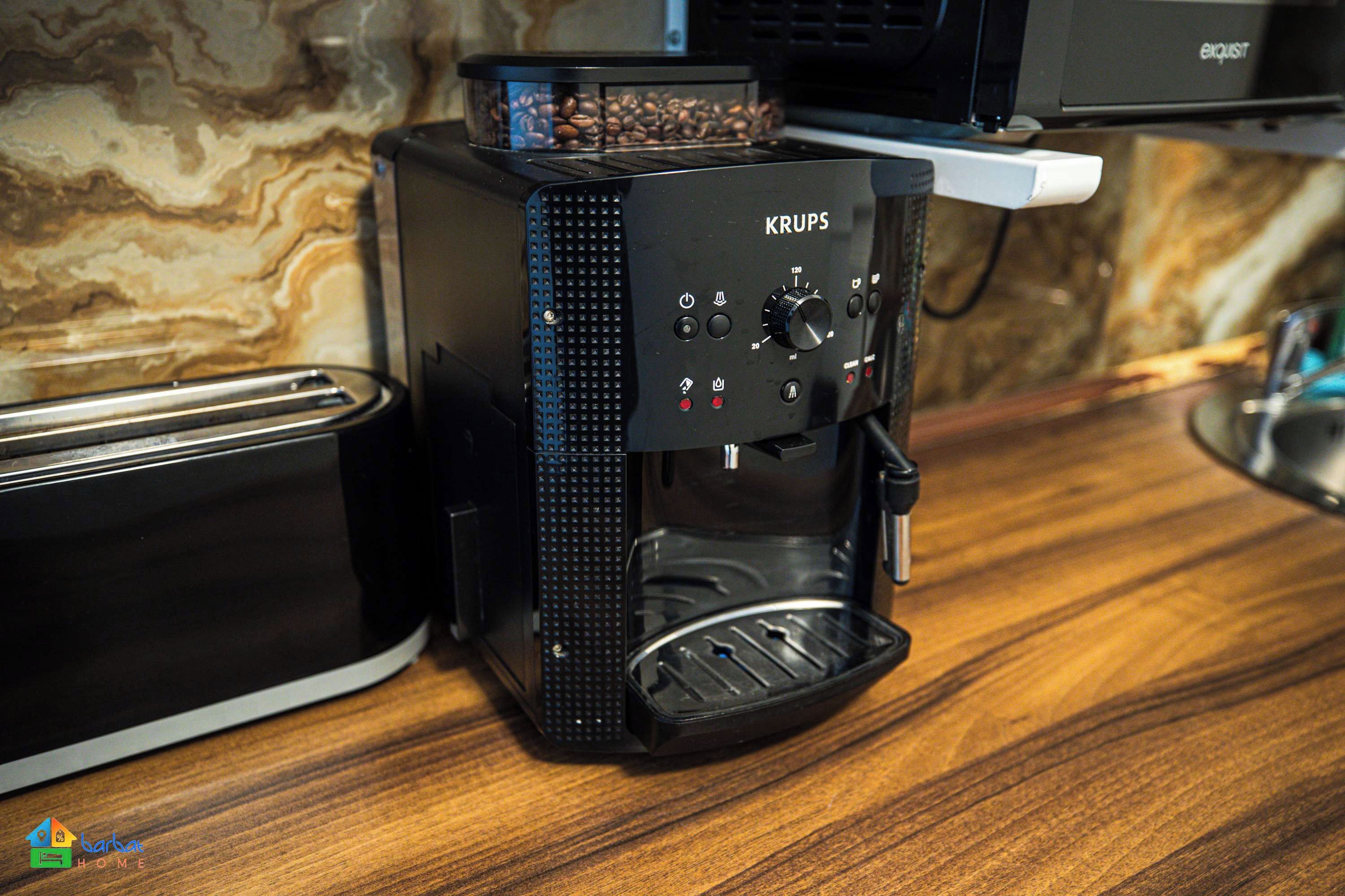Eine KRUPS Kaffeemaschine steht auf einer Holz Arbeitsplatte, zusammen mit einem Toaster. Im Hintergrund ist eine Wand mit einem marmorähnlichen Muster zu sehen.