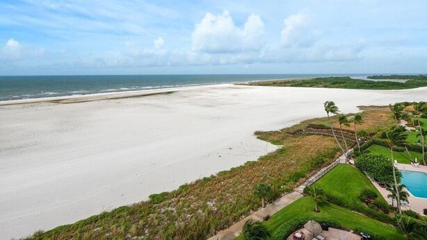 A beach view with a clear sky, a sandy beach, and a pool area with a fence and palm trees. - Beachfront 1BR condo on Marco Island with pool, WiFi, and kitchen (Marco Island)