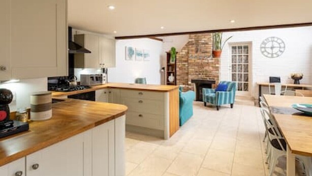A modern kitchen with white cabinetry, a wooden countertop, and a dining area with a blue chair and a table. - Charming Oxford Cottage (Oxfordshire)