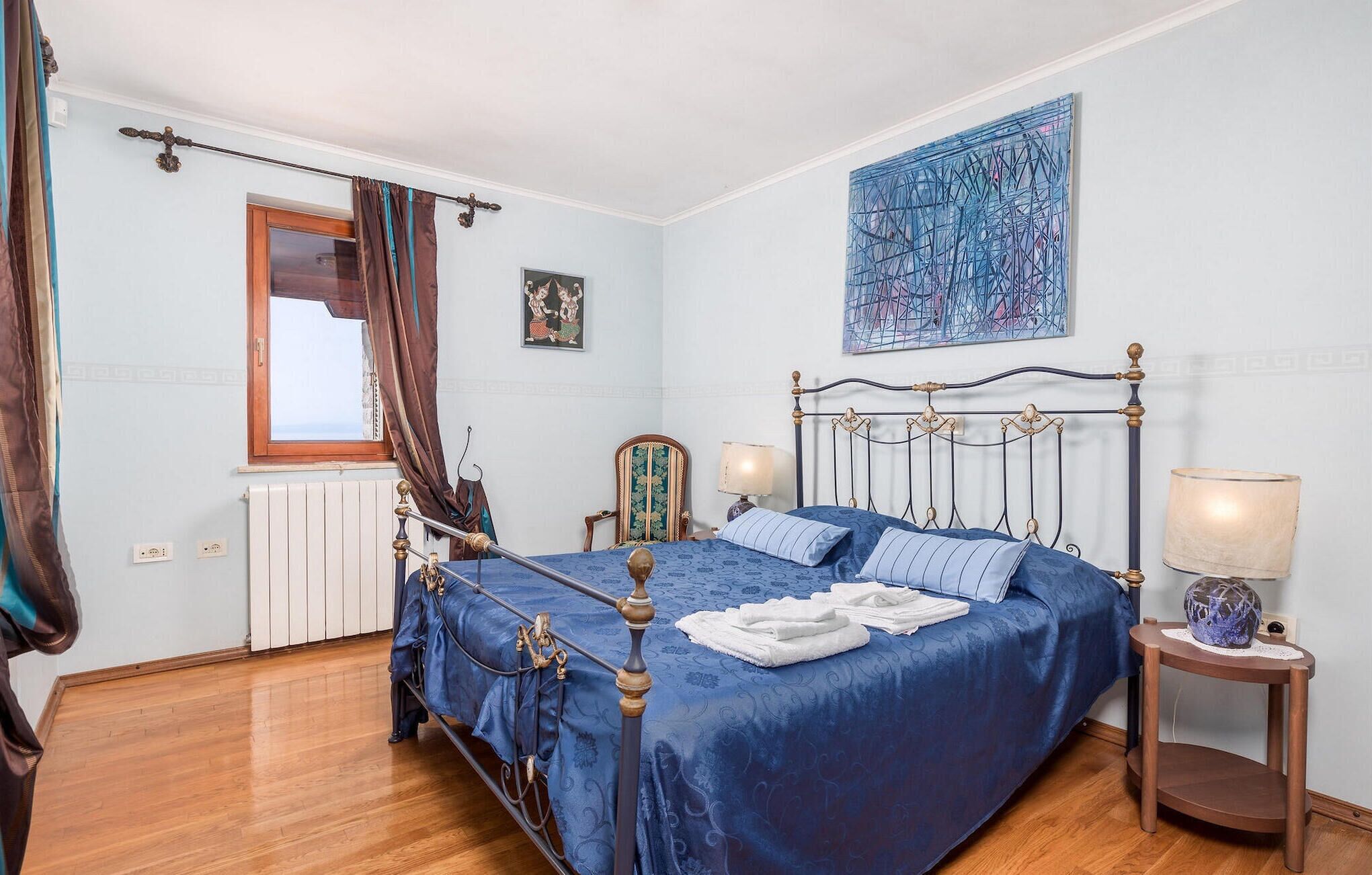 Une chambre à coucher avec un lit, deux tables de chevet, une fenêtre, un radiateur et un tableau au mur.