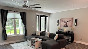 A modern living room with a sectional sofa, a coffee table, a fireplace, and a large window with curtains. - Hidden Hollow - Your Outdoor destination (Greig)
