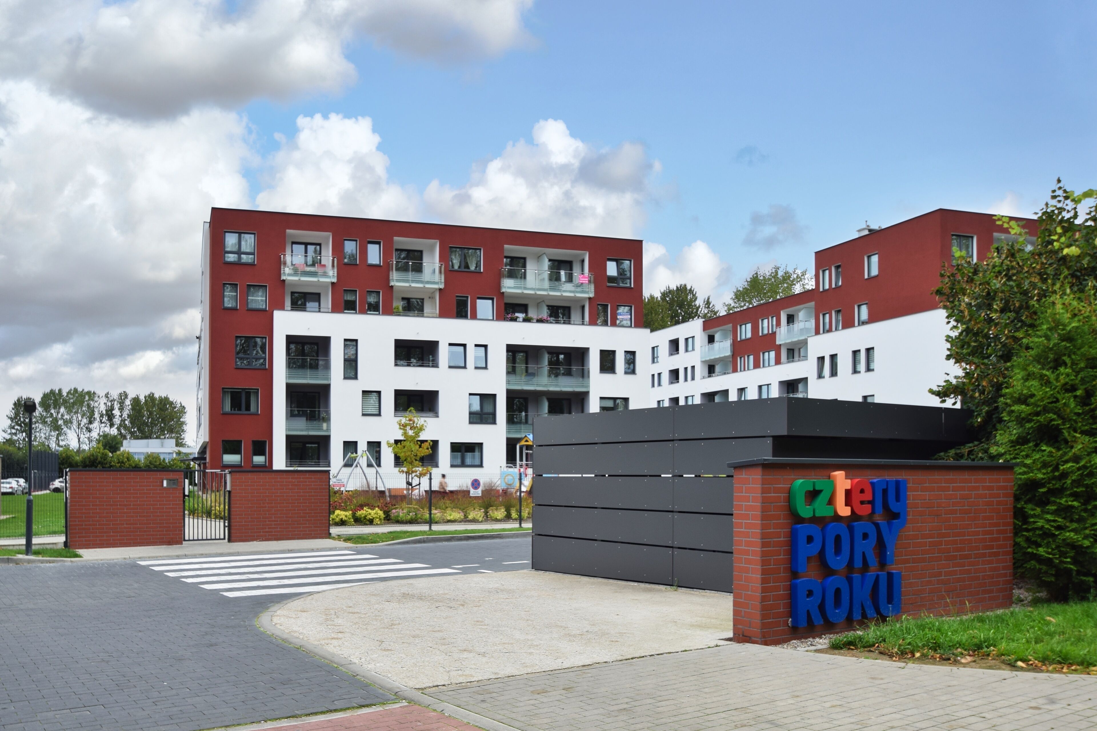 A multi-story building with a red and white facade, a sign reading "Cztery Pory Roku," and a gated entrance.