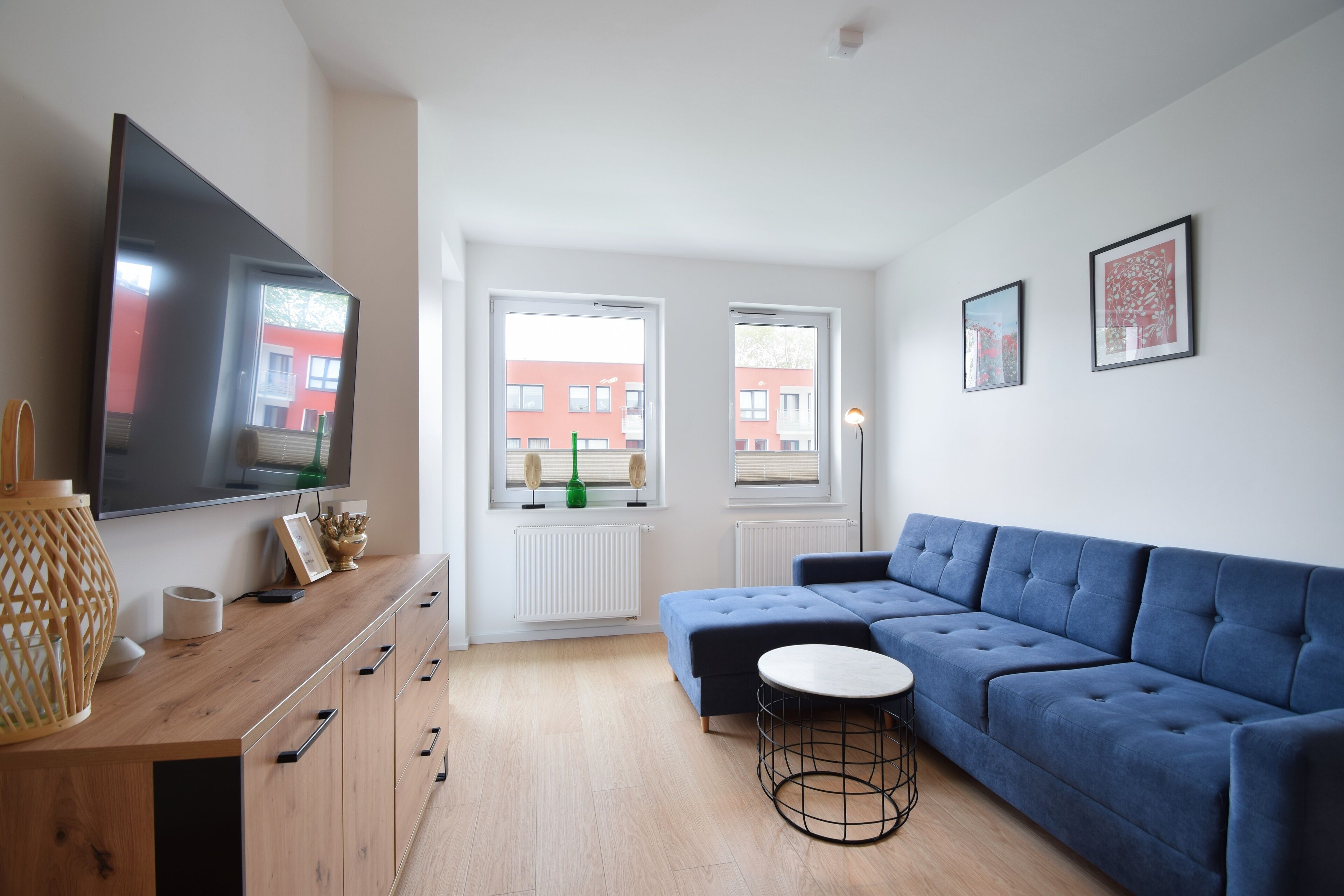 A modern living room with a large flat-screen TV, a blue sectional sofa, a small round table, and a wooden cabinet.