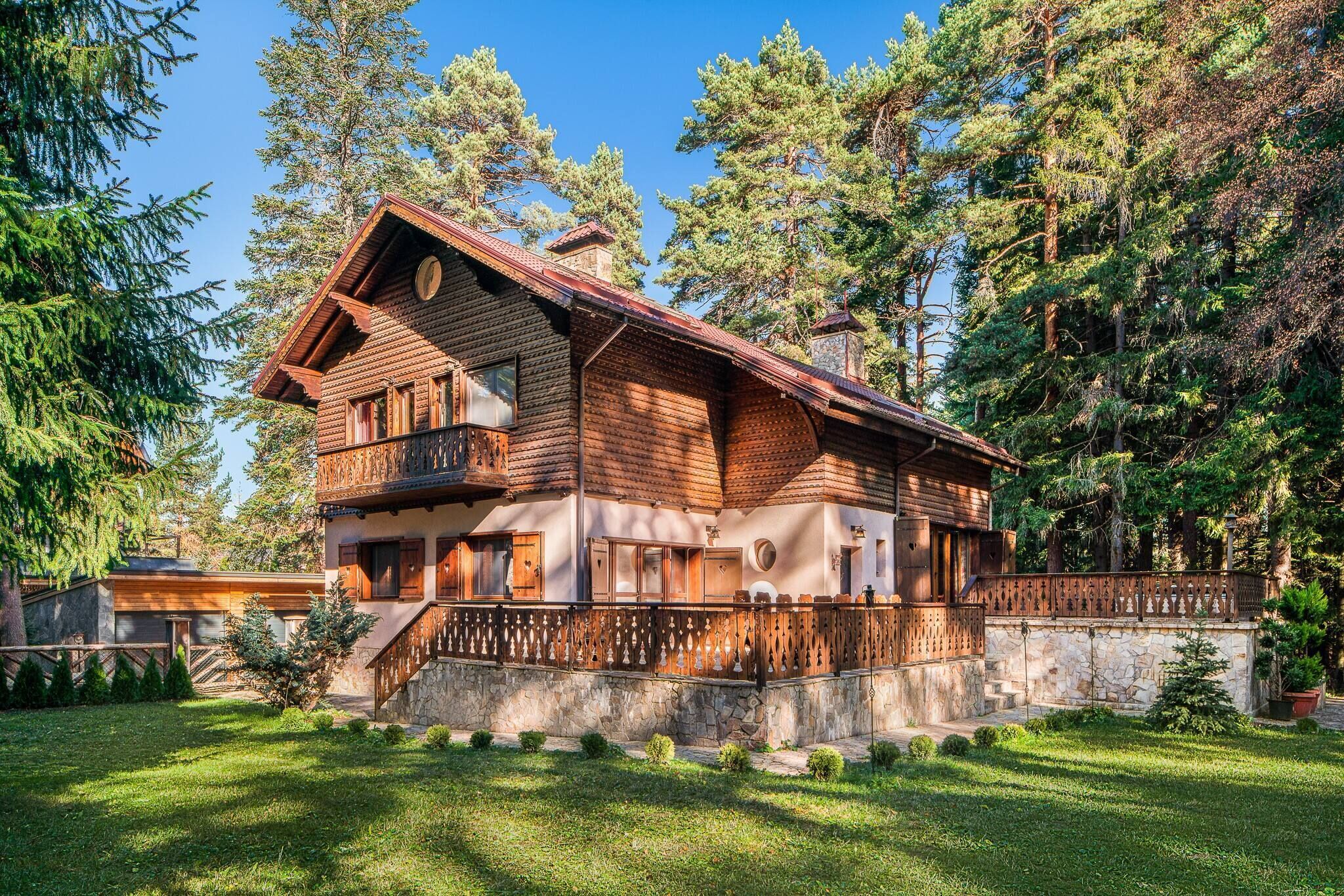 Une cabane en rondins à deux étages, dotée d’un balcon, entourée d’un mur de pierre et d’une végétation luxuriante.