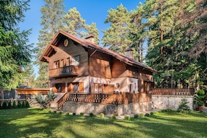 A two-story log cabin with a balcony, surrounded by a stone wall and lush greenery.