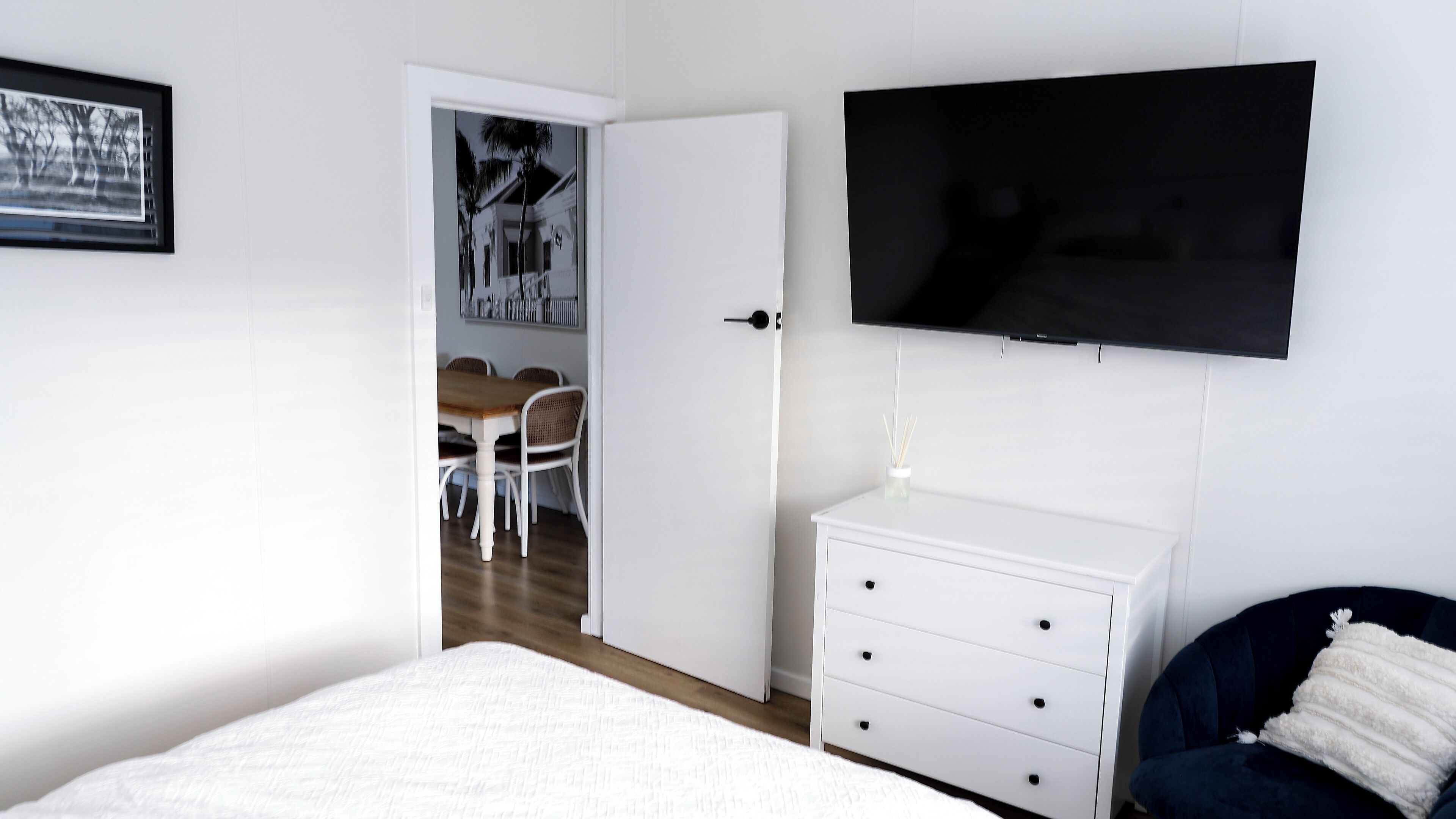A bedroom with a bed, a dresser, a TV, and a dining area visible through an open door.