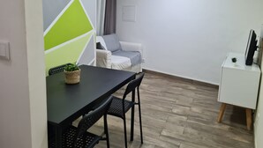 A small, modern living space with a dining area featuring a black table and chairs, a grey sofa, and a white cabinet with a TV.