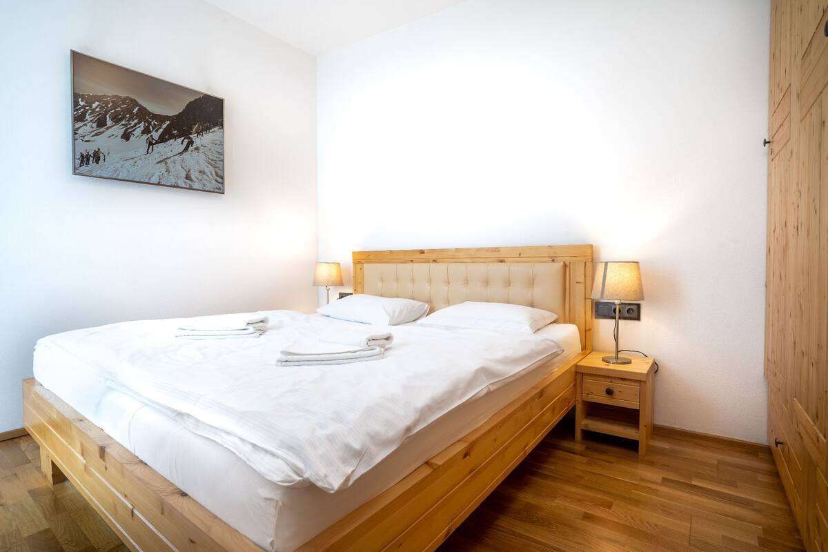 A neatly made bed with white linens, a wooden headboard, and two bedside lamps.