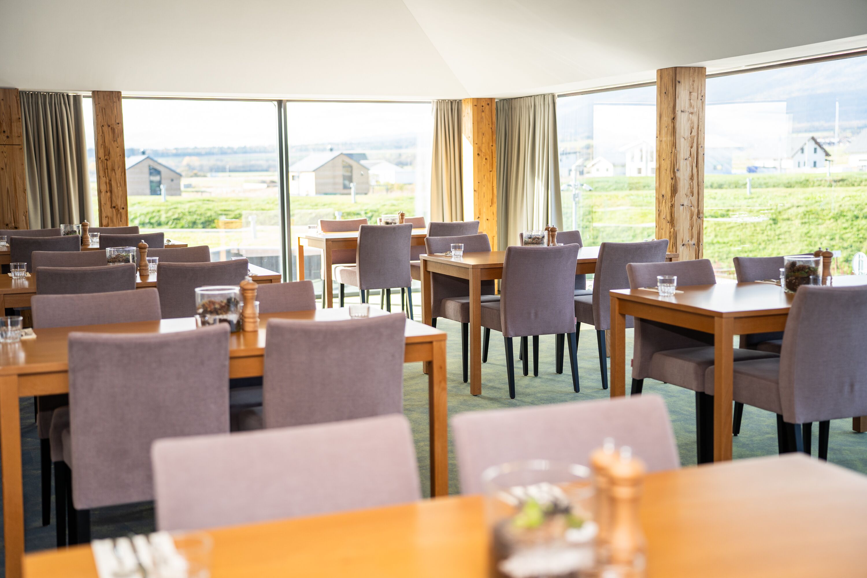 A restaurant with multiple tables and chairs, large windows with curtains, and a view of the countryside.