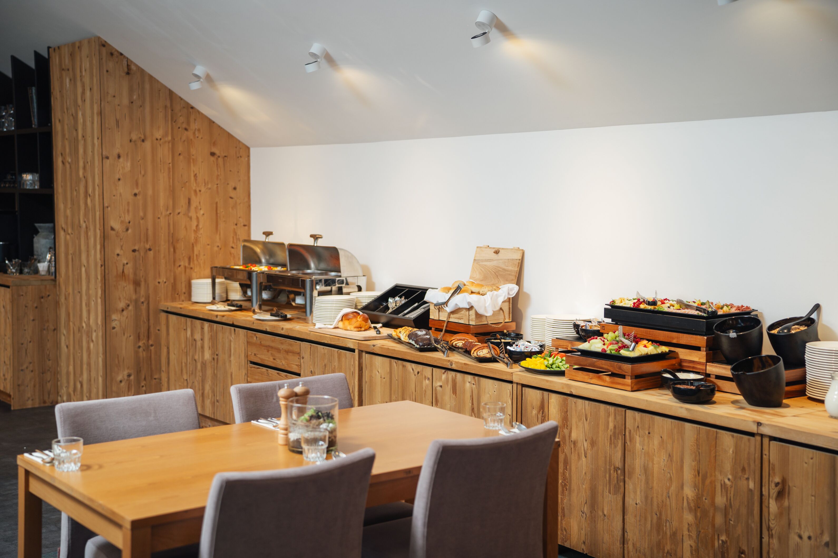 Un comedor moderno con paneles de madera, un mostrador de buffet con diversas opciones de comida y un área de estar con mesas y sillas.