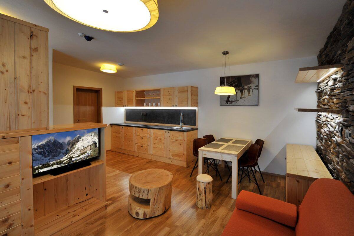 A modern kitchen with wooden cabinets, a dining area with a table and chairs, a television mounted on a wooden stand, and a stone accent wall.