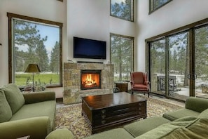 A living room with a stone fireplace, a flat-screen TV, green sofas, and large windows.