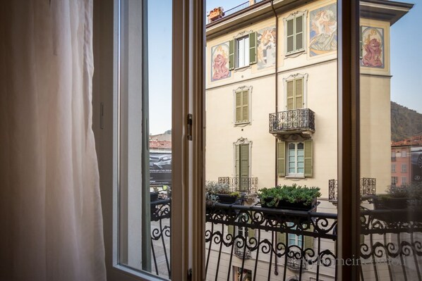 A view through a window with a balcony, featuring a building with green shutters and decorative artwork.