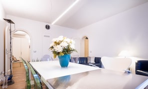A modern hotel room with a white table, a blue vase with flowers, a clock on the wall, and a lamp.