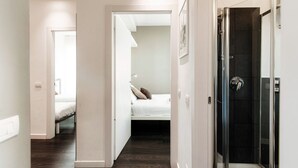 Quarto de hotel moderno com chuveiro de vidro, cama com roupa de cama branca e um pequeno espaço de estar com uma cadeira e mesa.